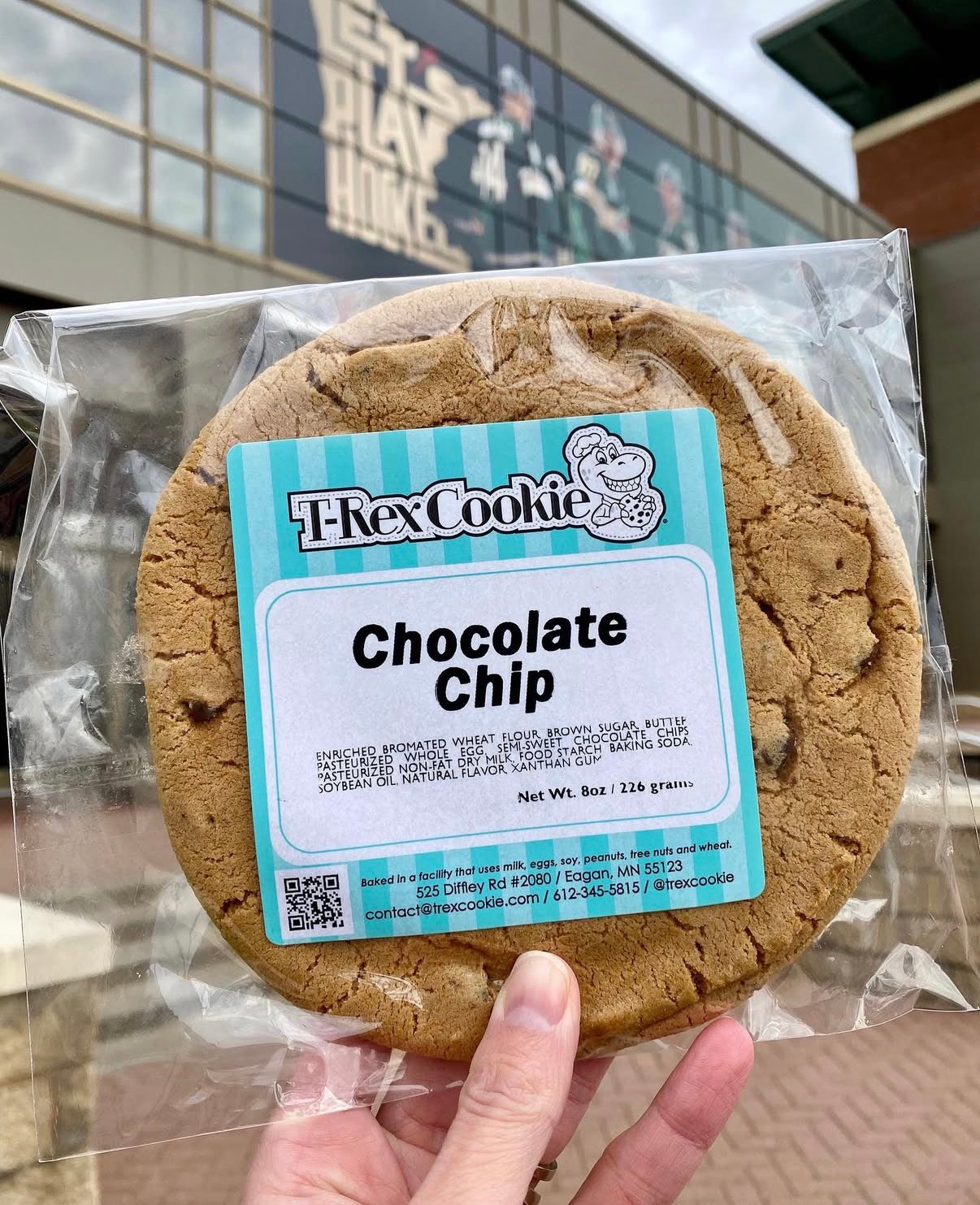 Chocolate chip cookie with a 'T-Rex Cookie' label held in front of a building.