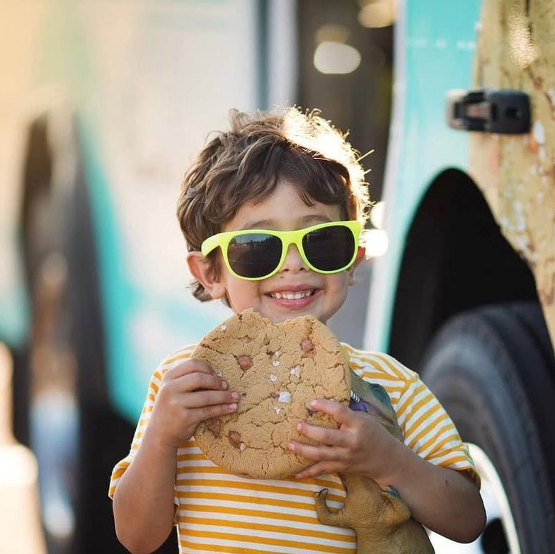 Child wearing sunglasses holding a cookie in front of a teal bus.